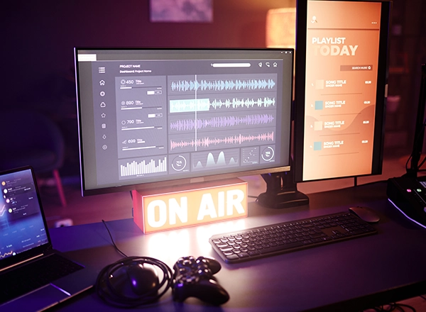 A professional broadcasting workstation featuring dual monitors with audio editing software, a lit "ON AIR" sign, a laptop, and a gamepad controller.