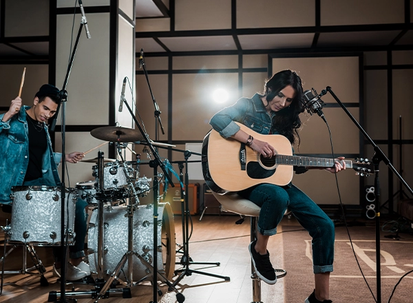 A musician playing an acoustic guitar while a drummer plays along in a recording studio.