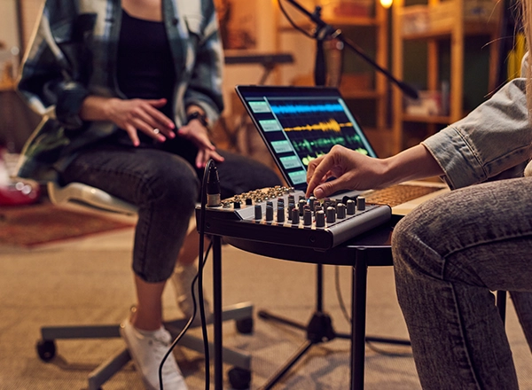 A close-up of a person adjusting an audio mixing console connected to a laptop running sound editing software during a recording session.