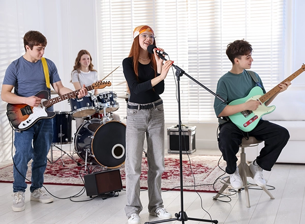 A teenage band rehearsing in a bright room, featuring a young female vocalist, an electric guitarist, and a female drummer.