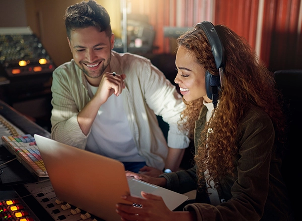 Two music professionals smiling and collaborating over a laptop inside a recording studio control room.