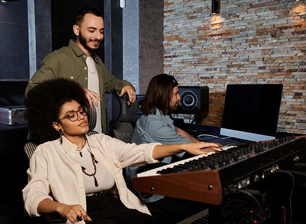 Three music producers collaborating on a track around a synthesizer keyboard in a recording studio.