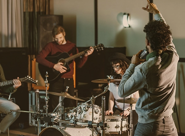 An energetic four-piece band practicing in a rehearsal studio, with a lead singer facing away in the foreground raising his hand.