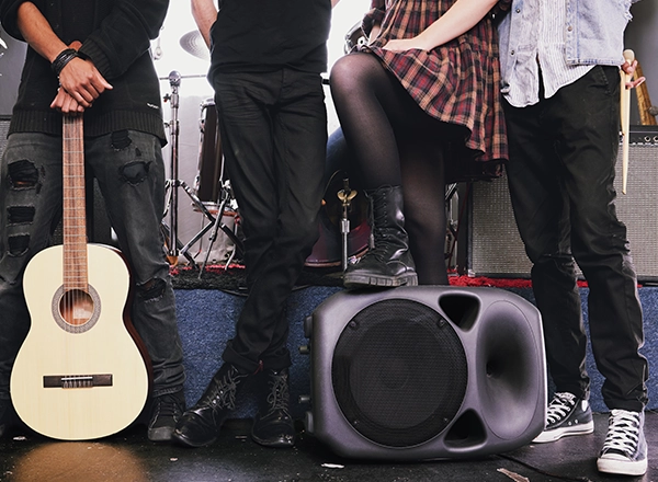 A cropped shot from the waist down of a four-piece band posing on a stage with an acoustic guitar, drumsticks, and a speaker monitor.