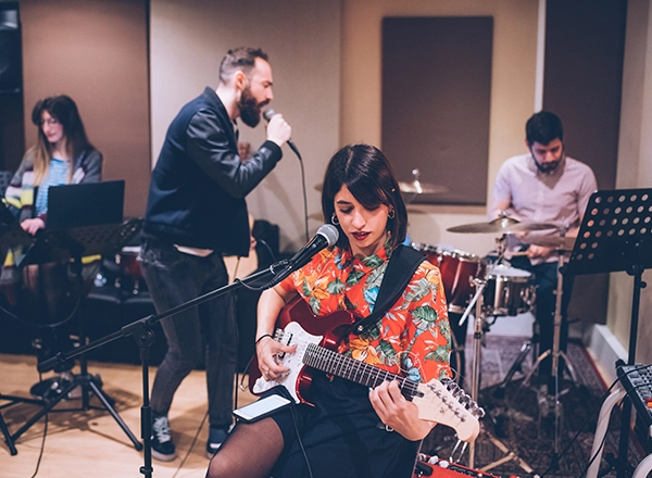 A four-piece indie band practicing in a rehearsal studio, featuring a woman playing a red electric guitar in the foreground.